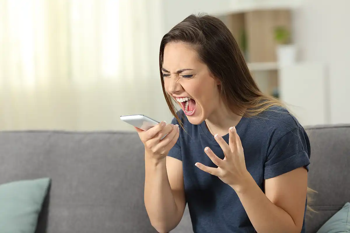 A woman yelling into her smartphone while sitting on a couch after not protecting her company with Defensive Domain Registration.